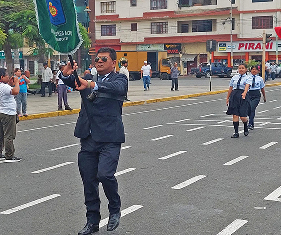Portaestandarte con terno oscuro y lentes conduce la marcha del colegio por una avenida de Chimbote; detrás, escolares con blusa blanca, corbata y falda azul marino.