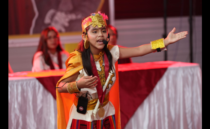 Escolar con vestido de gala andino y micrófono de diadema declama en un escenario con mantas rojas y blancas durante un concurso escolar.