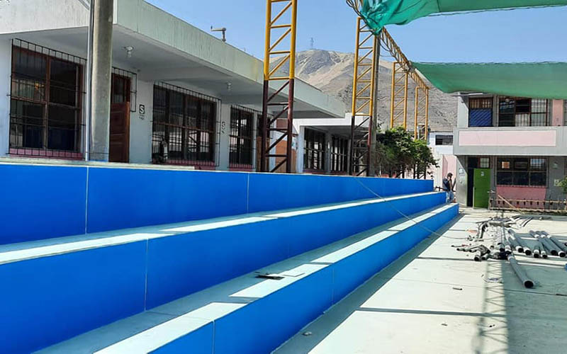 Graderías azules nuevas de la losa deportiva, torres amarillas de la cobertura metálica y pabellones blancos del colegio con el cerro de Coishco al fondo.