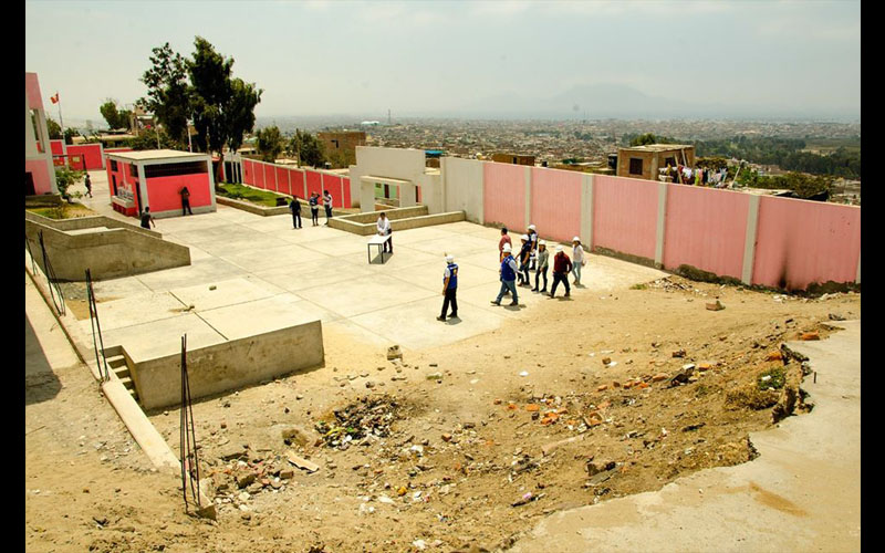 Patio de cemento con escombros y terreno abierto al fondo, cerco rosado a la derecha y pabellón crema de dos pisos a la izquierda; unos escolares caminan por la losa.