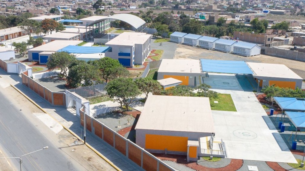 Vista aérea del colegio con pabellones de acentos naranja y celeste, losa con arcos pintados, cobertura arqueada y cerco perimétrico blanco y ladrillo.