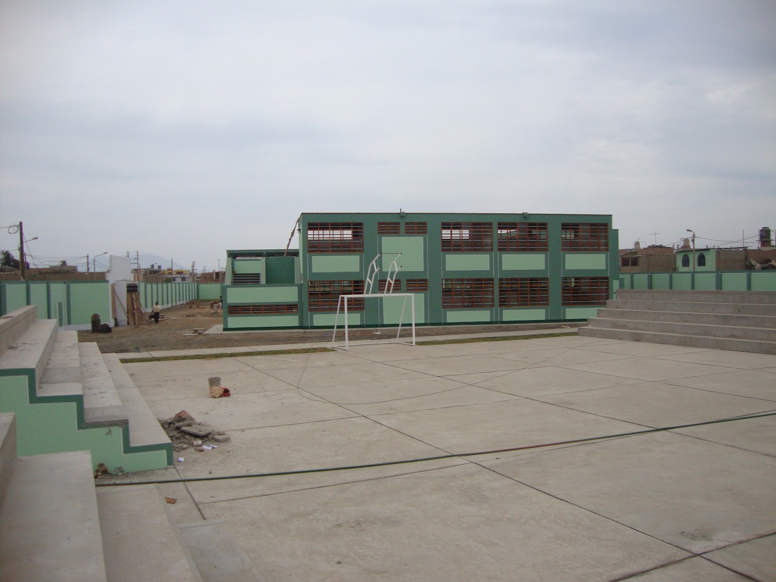 Loza deportiva de cemento con tribunas de concreto y arcos blancos, frente al pabellón de dos pisos pintado de verde.