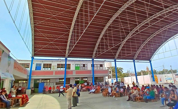 Patio del colegio cubierto por el arco metálico rojizo del cobertor sobre columnas azules; al fondo el pabellón crema y salmón de dos pisos y la comunidad reunida en la ceremonia.