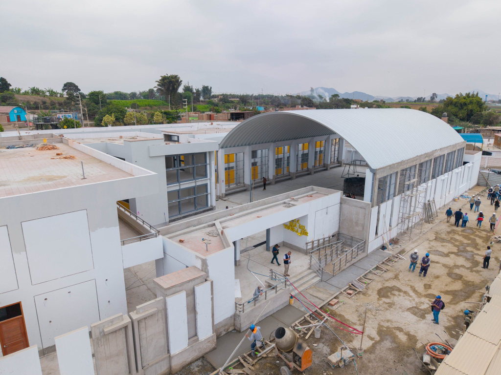 Vista aérea de la obra del colegio de El Castillo: pabellones blancos de dos niveles, sala con techo metálico en arco, trabajadores con casco y mezcladora en el patio de tierra.