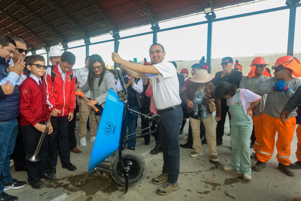Autoridades y escolares con casaca roja durante el acto de inicio de obra; un voluntario vuelca cemento desde una carretilla azul.