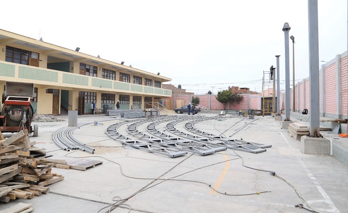 Patio del colegio con la estructura metálica curva de la cobertura desarmada sobre la losa, pabellón crema de dos pisos a la izquierda y cerco rosado al fondo.