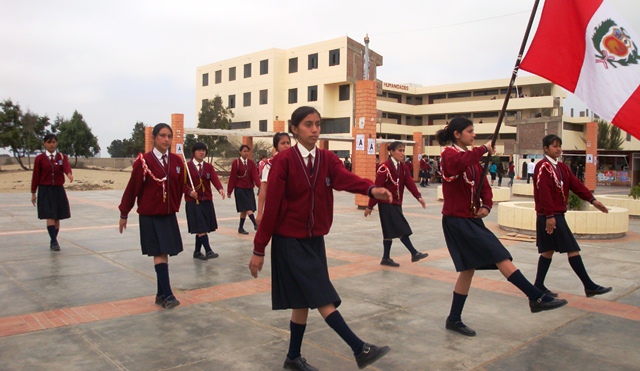 Estudiantes de uniforme guindo desfilan con la bandera peruana frente al pabellón crema del colegio experimental de la UNS.